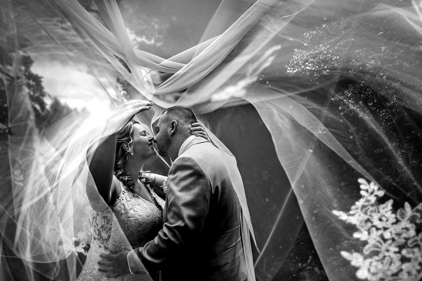 Zwart-witte bruiloftsfoto: bruid en bruidegom kussen onder een sluier, met bloemen en lichtvlekken.