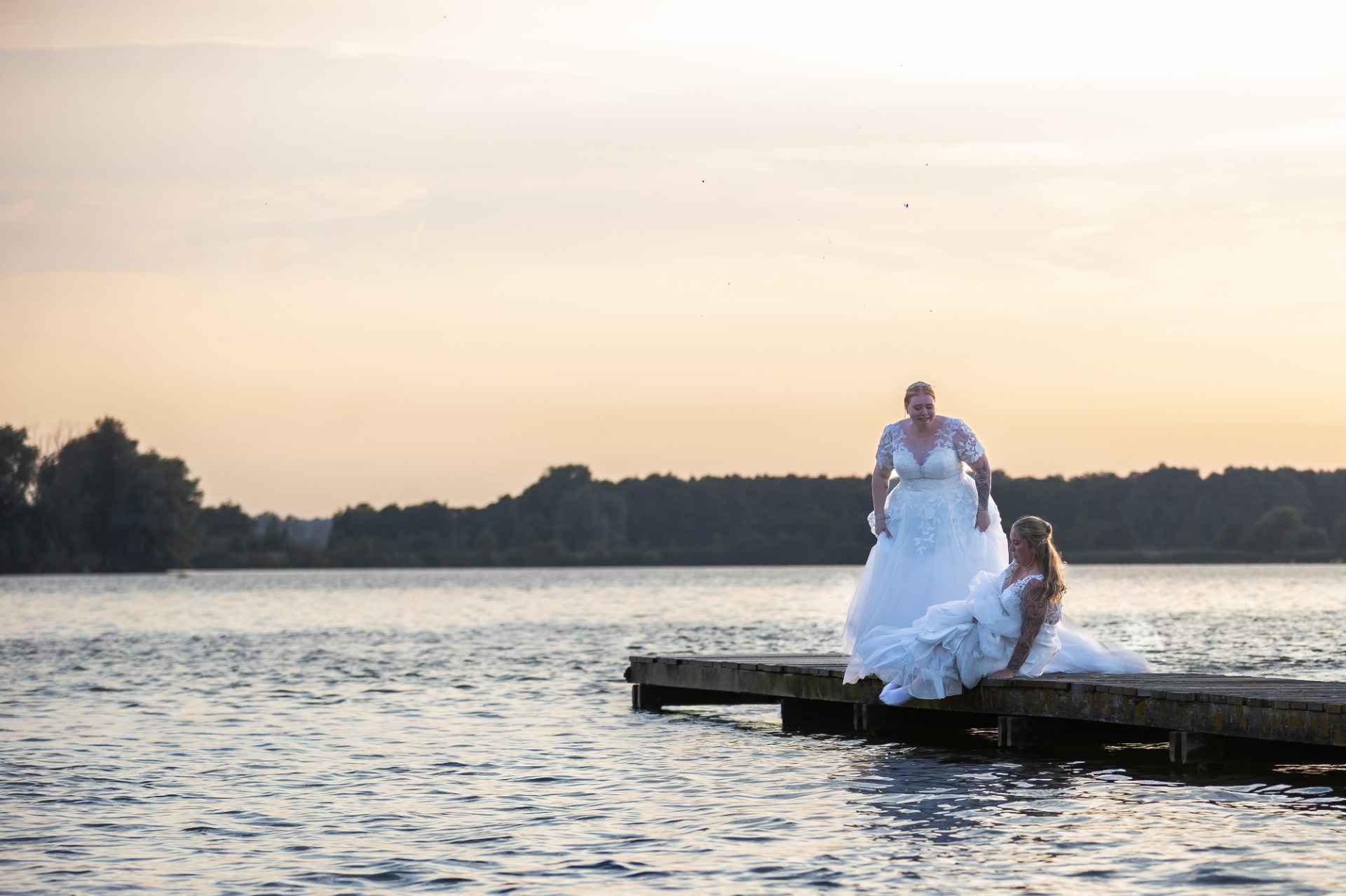 Bruidsmeisjes in witte jurken zitten op een houten steiger bij een meer tijdens zonsondergang.
