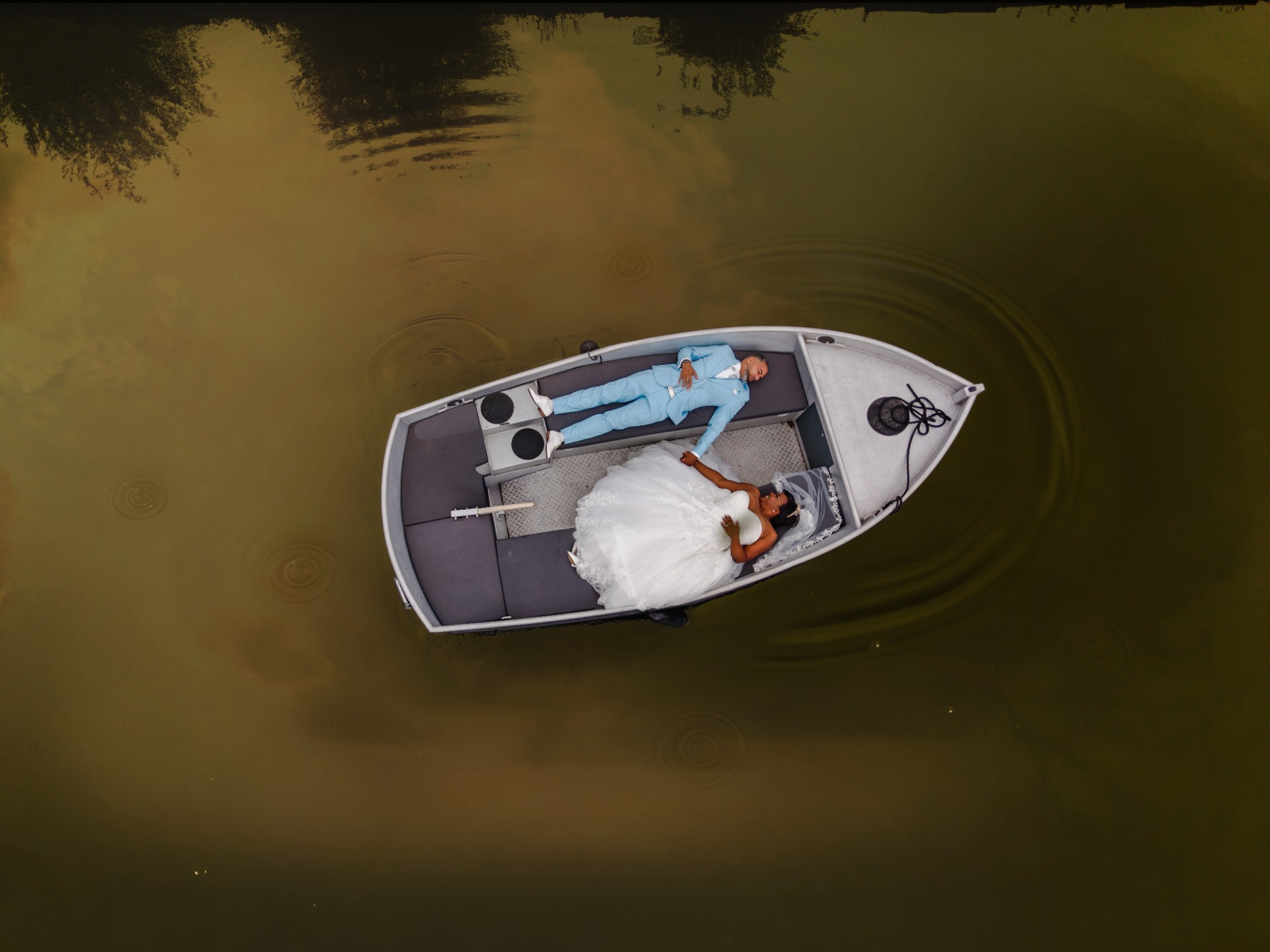 Twee personen liggen in een kleine boot op een meer, met witte trouwjurk en blauwe outfit.